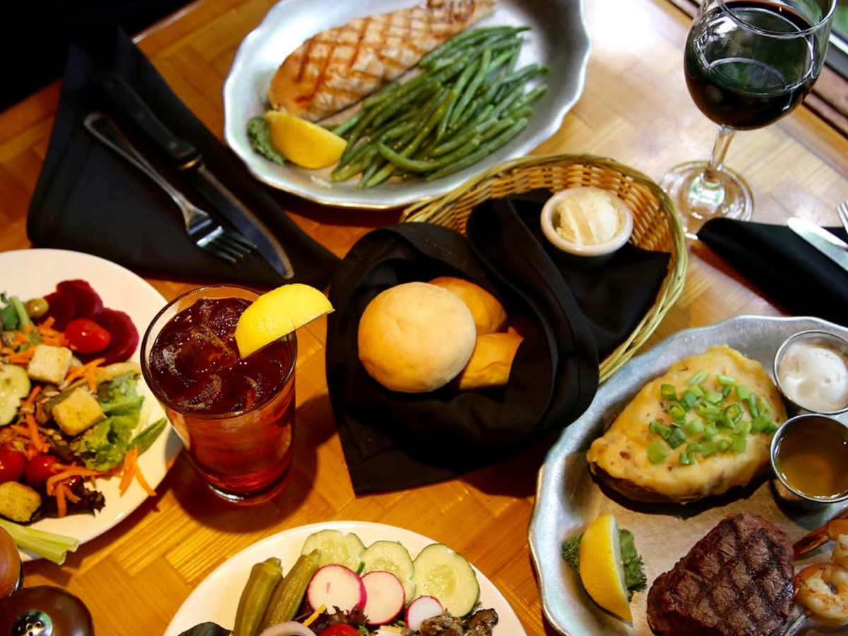 A hearty dinner spread featuring grilled chicken with green beans, a filet with shrimp and loaded baked potato, fresh garden salad, assorted pickled vegetables, dinner rolls with butter, and drinks including iced tea and red wine.
