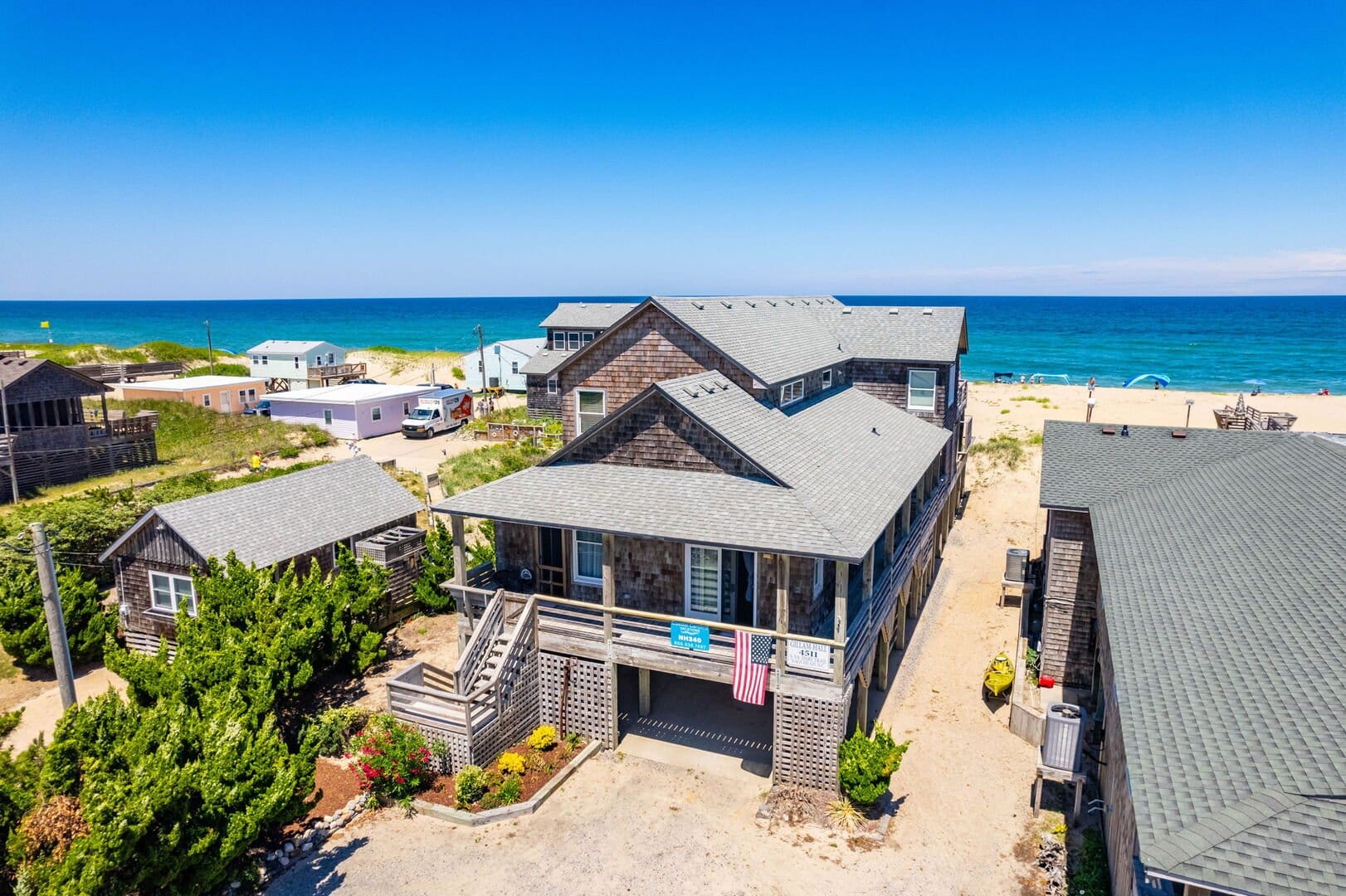 Elevated gray shingle-style beach house with wraparound porch and ocean view, located steps from a sandy shoreline with neighboring coastal homes.