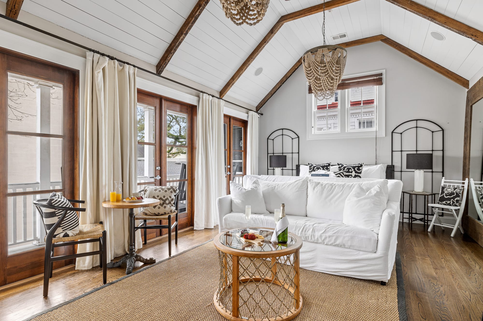 A charming Charleston-style bedroom with vaulted ceilings, wood beams, and soft white décor creates a calm and relaxing retreat. French doors open to a balcony, letting in fresh air and natural light.