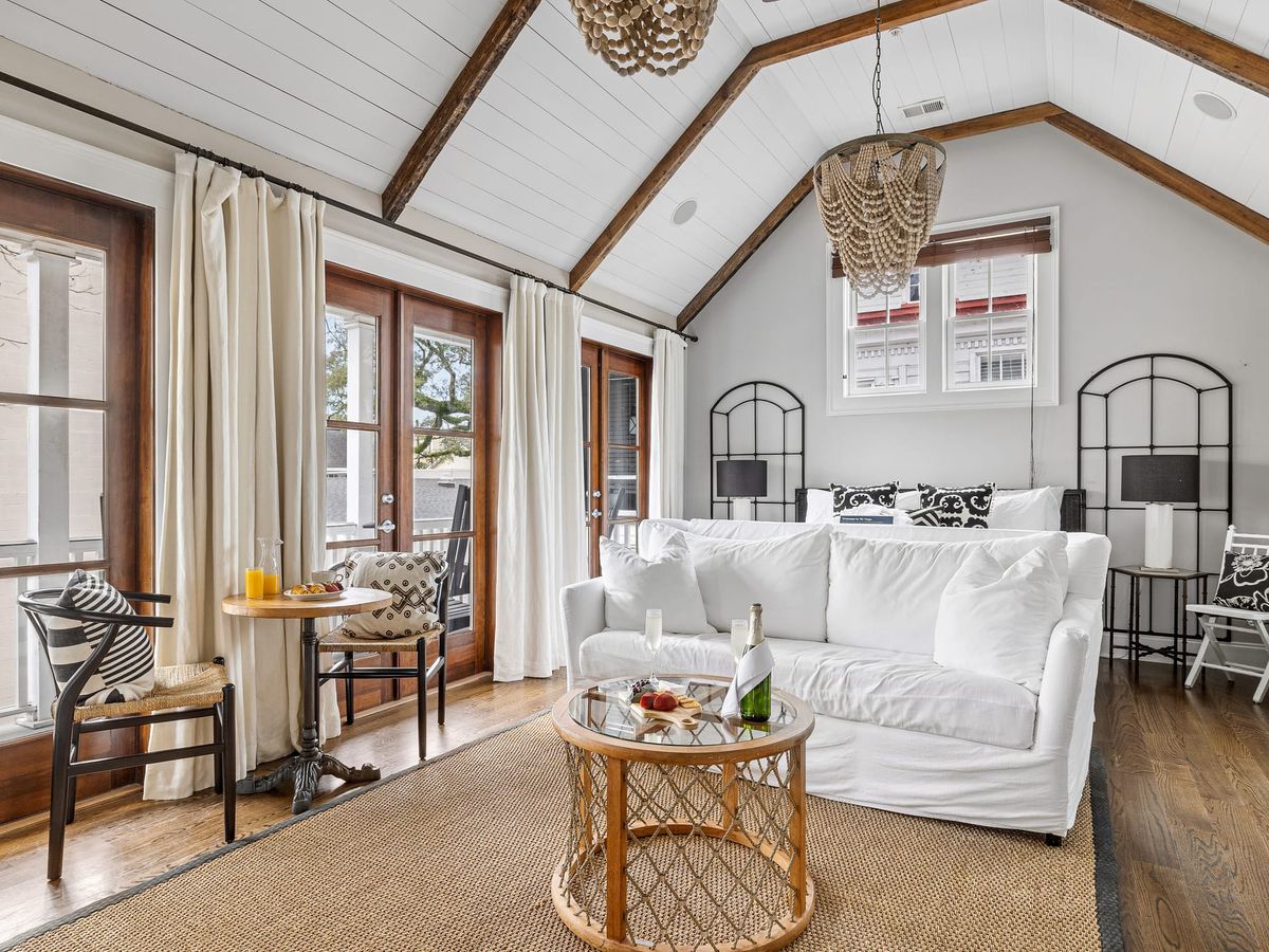 A charming Charleston-style bedroom with vaulted ceilings, wood beams, and soft white décor creates a calm and relaxing retreat. French doors open to a balcony, letting in fresh air and natural light.