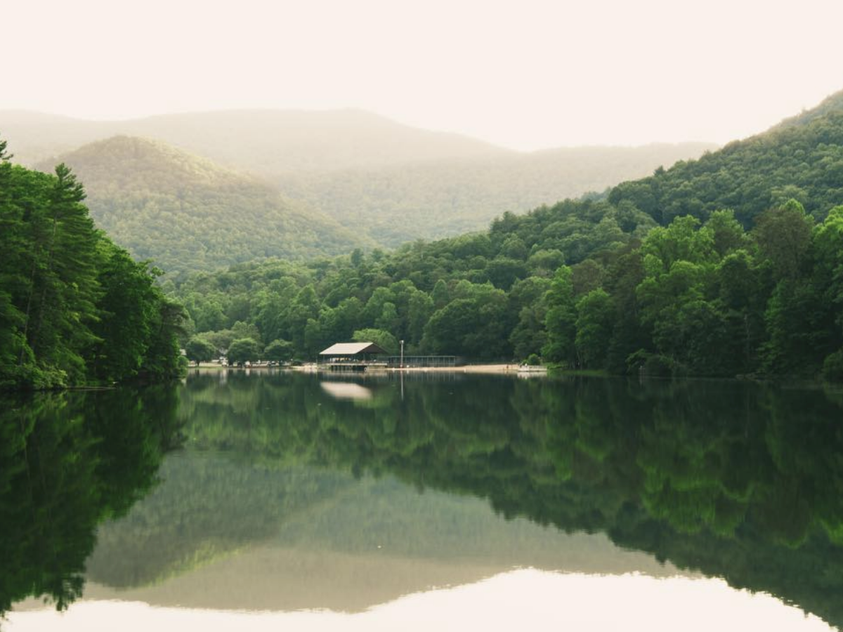 A peaceful lake surrounded by lush green mountains reflects the trees and a small dock with a shelter under a hazy sky.