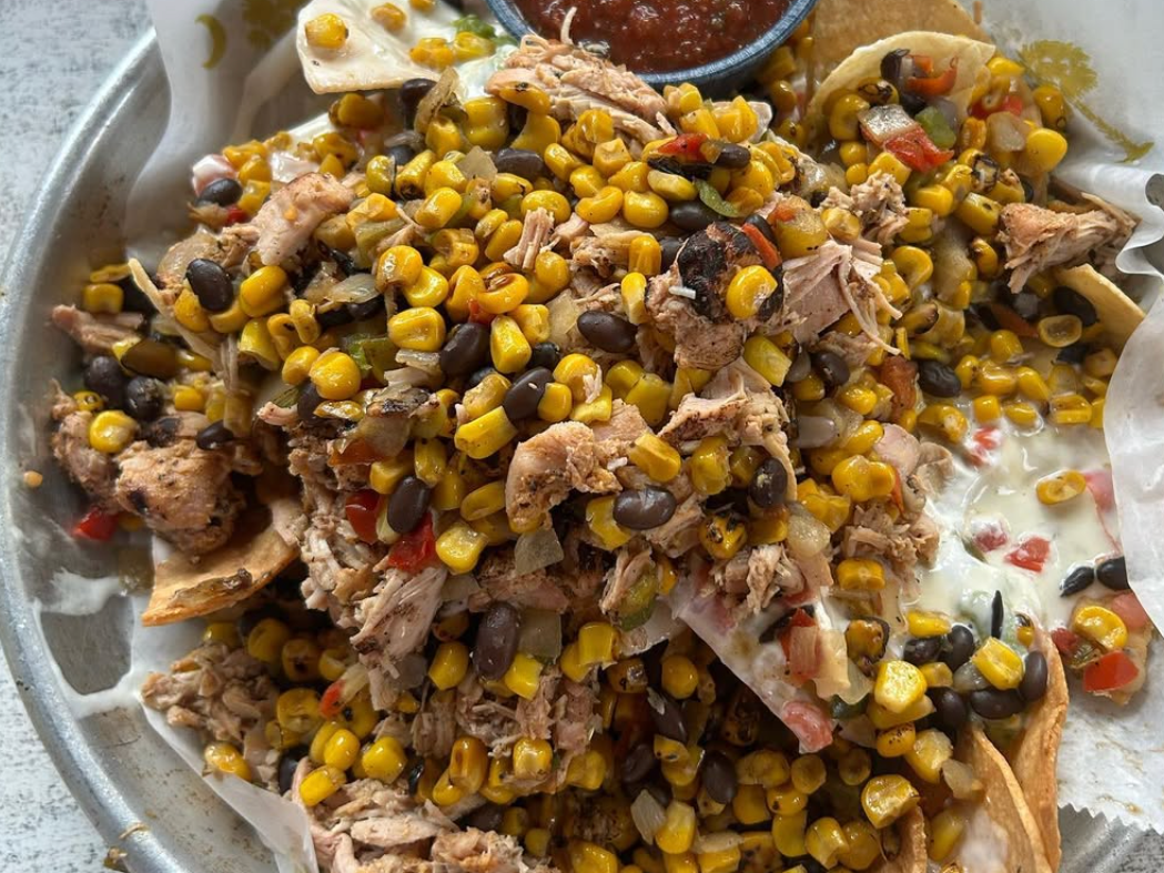 A tray of loaded nachos topped with pulled chicken, roasted corn, black beans, diced peppers, and melted cheese, served with a side of salsa.
