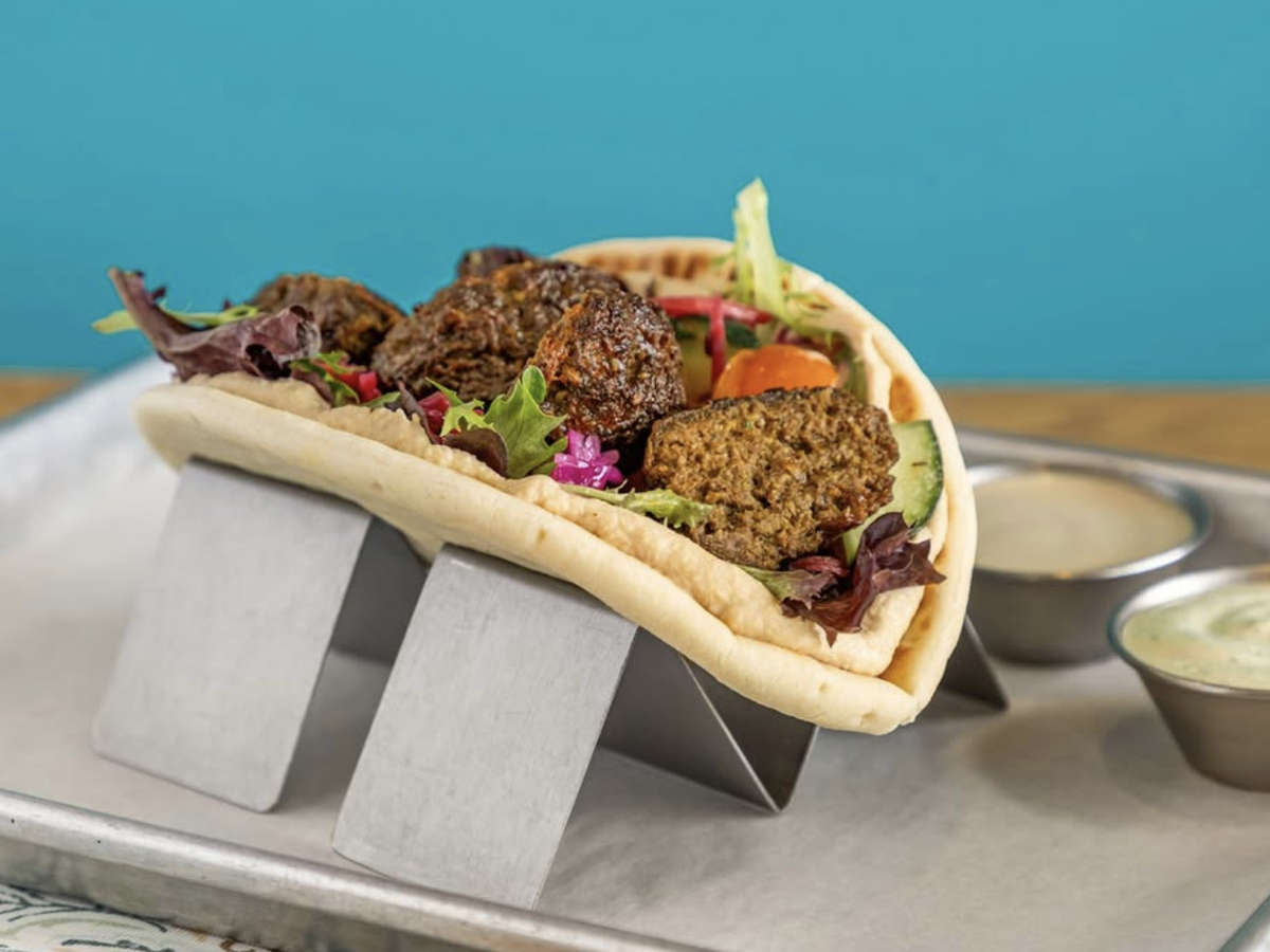 A fluffy pita is loaded with crispy falafel, mixed greens, cucumbers, pickled vegetables, and served with a side of creamy dipping sauces on a metal taco rack.