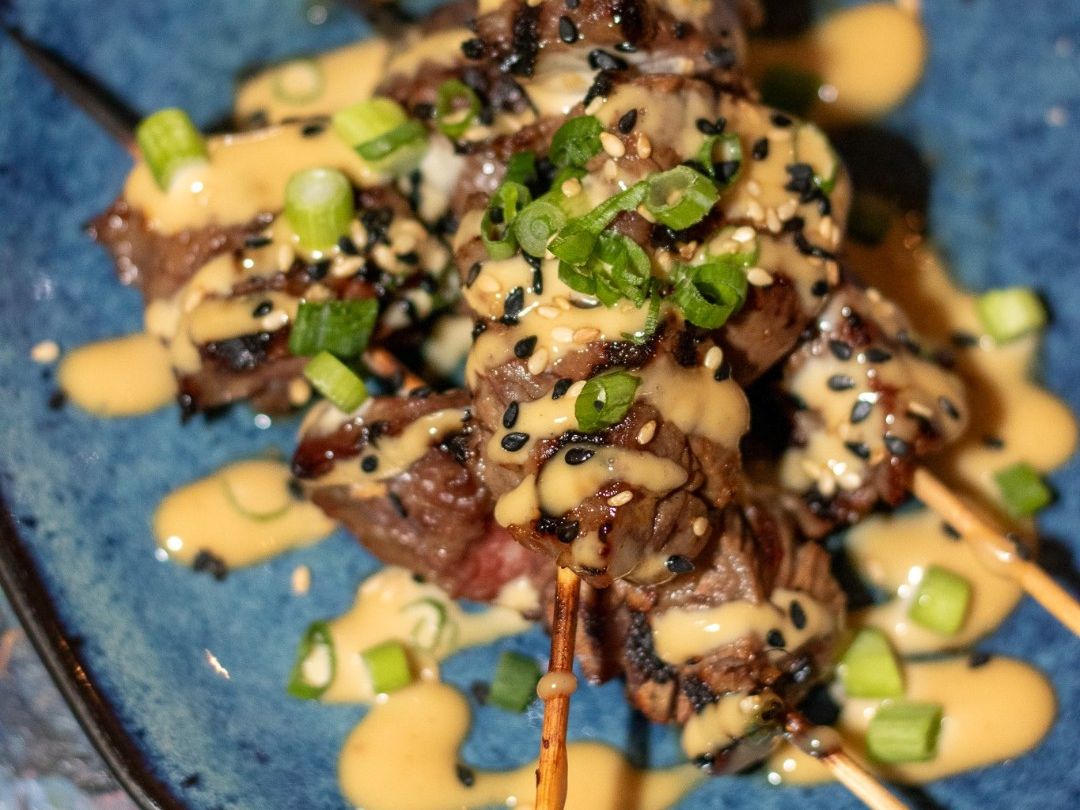 Juicy grilled beef skewers drizzled with creamy sauce, sesame seeds, and scallions served on a blue plate at Café Tu Tu Tango in Orlando. A perfect mix of smoky, savory, and fresh flavors.