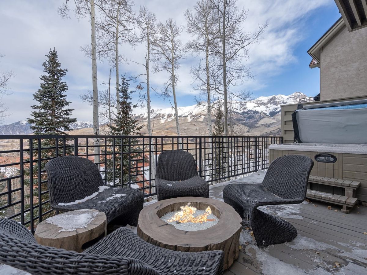 Mountain deck with wicker chairs around a fire pit, hot tub nearby, and snowy peaks visible through tall trees.