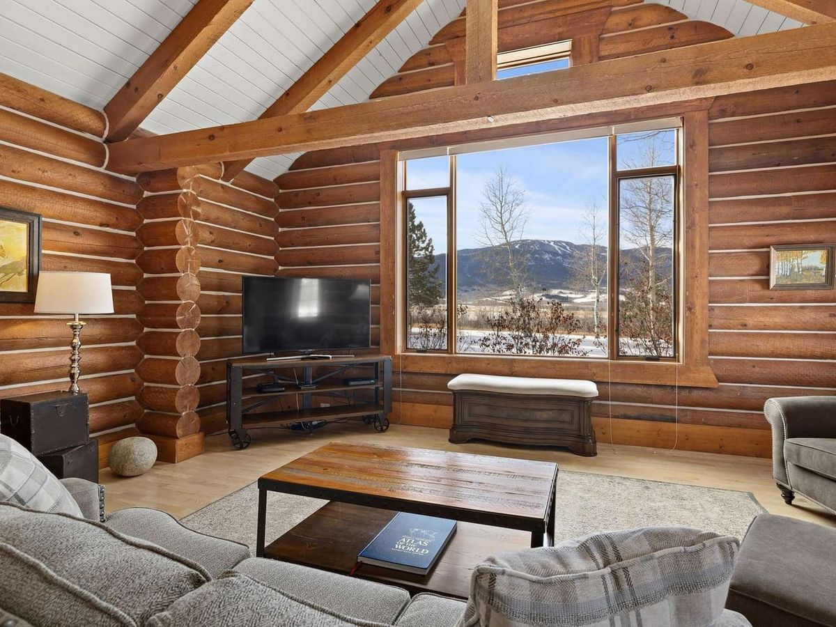A classic log cabin living room with soaring ceilings and big mountain views through tall windows. Warm wood beams and rustic charm make this a perfect escape after a day of hiking or skiing.