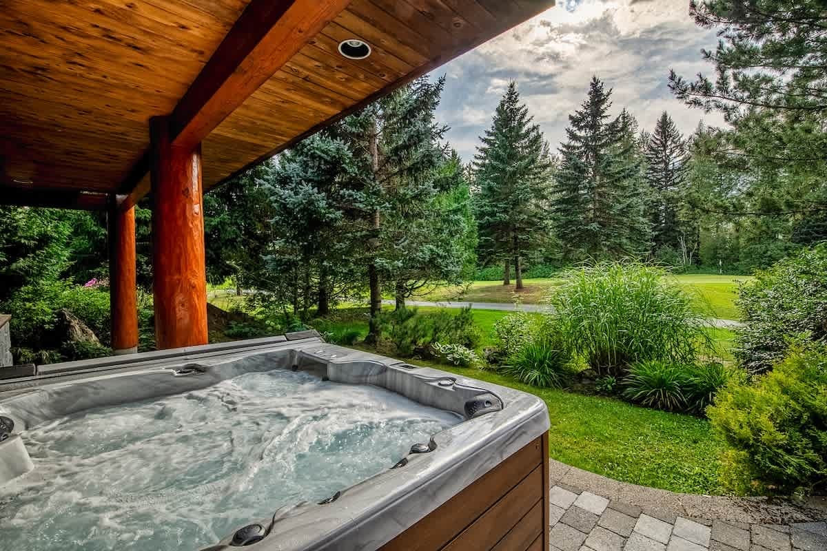 ubbling outdoor hot tub on a covered patio with wide views of green lawns, pine trees, and a peaceful mountain landscape. This private chalet spa setup creates a calm place to relax and enjoy the fresh air.