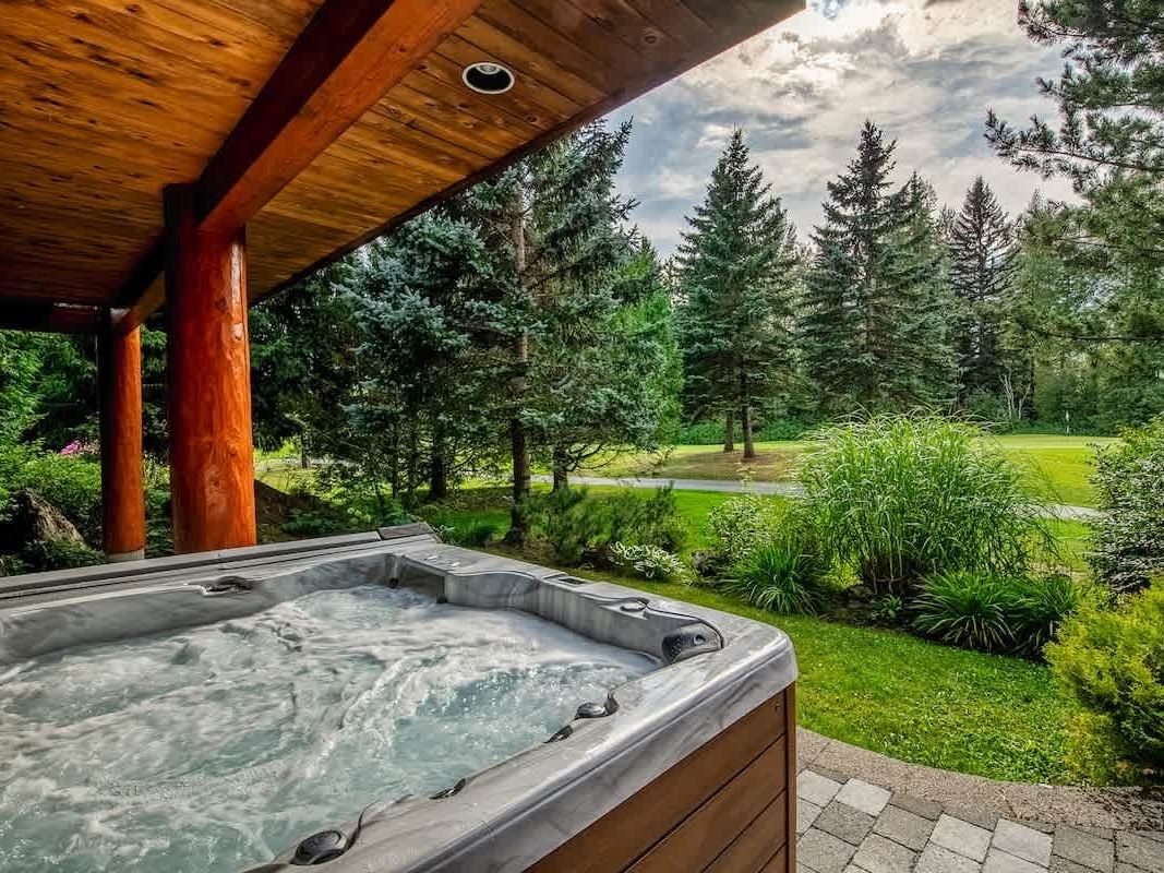 ubbling outdoor hot tub on a covered patio with wide views of green lawns, pine trees, and a peaceful mountain landscape. This private chalet spa setup creates a calm place to relax and enjoy the fresh air.