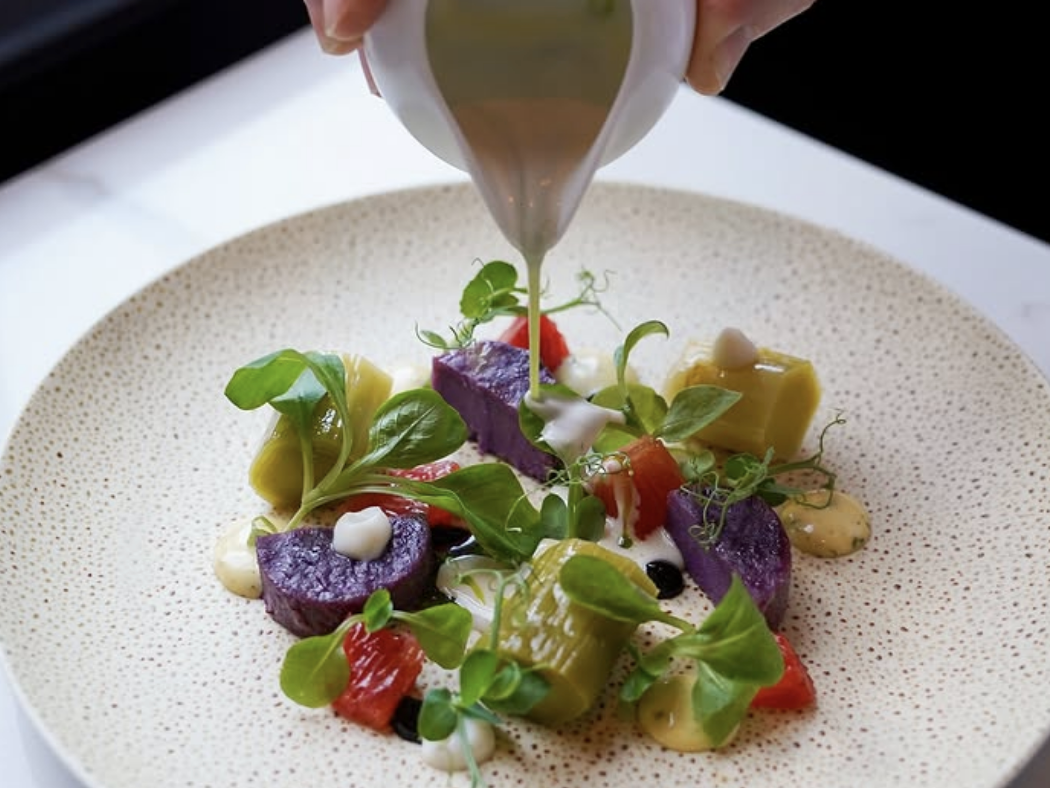 A hand pours dressing over a colorful gourmet dish featuring vibrant purple and yellow vegetables, fresh greens, and grapefruit segments, artfully plated with various sauces on a speckled ceramic plate.