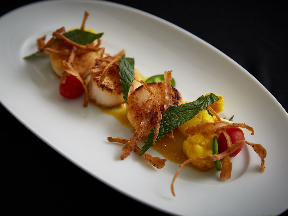 Seared scallops topped with crispy garnish and mint, served with cherry tomatoes and golden cauliflower. A fancy seafood dish perfect for a romantic dinner near the coast.