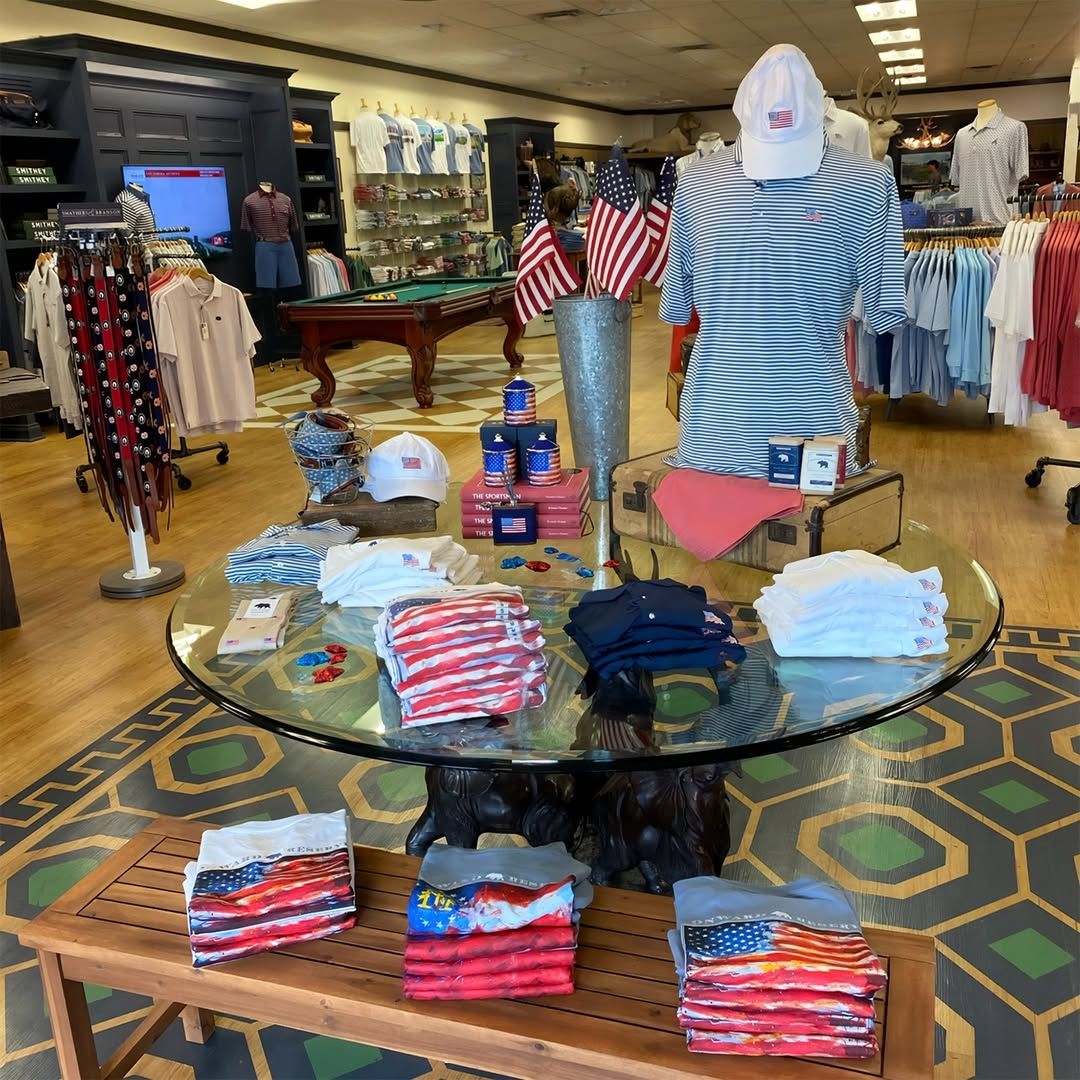 The interior of Onward Reserve displays a curated selection of American-themed apparel and accessories arranged on a round glass-top table with a carved bear base, surrounded by folded flag-print shirts, hats, and koozies. Polo shirts and belts line the walls and racks in the background, with a pool table and mounted deer head adding to the classic Southern sporting aesthetic. 