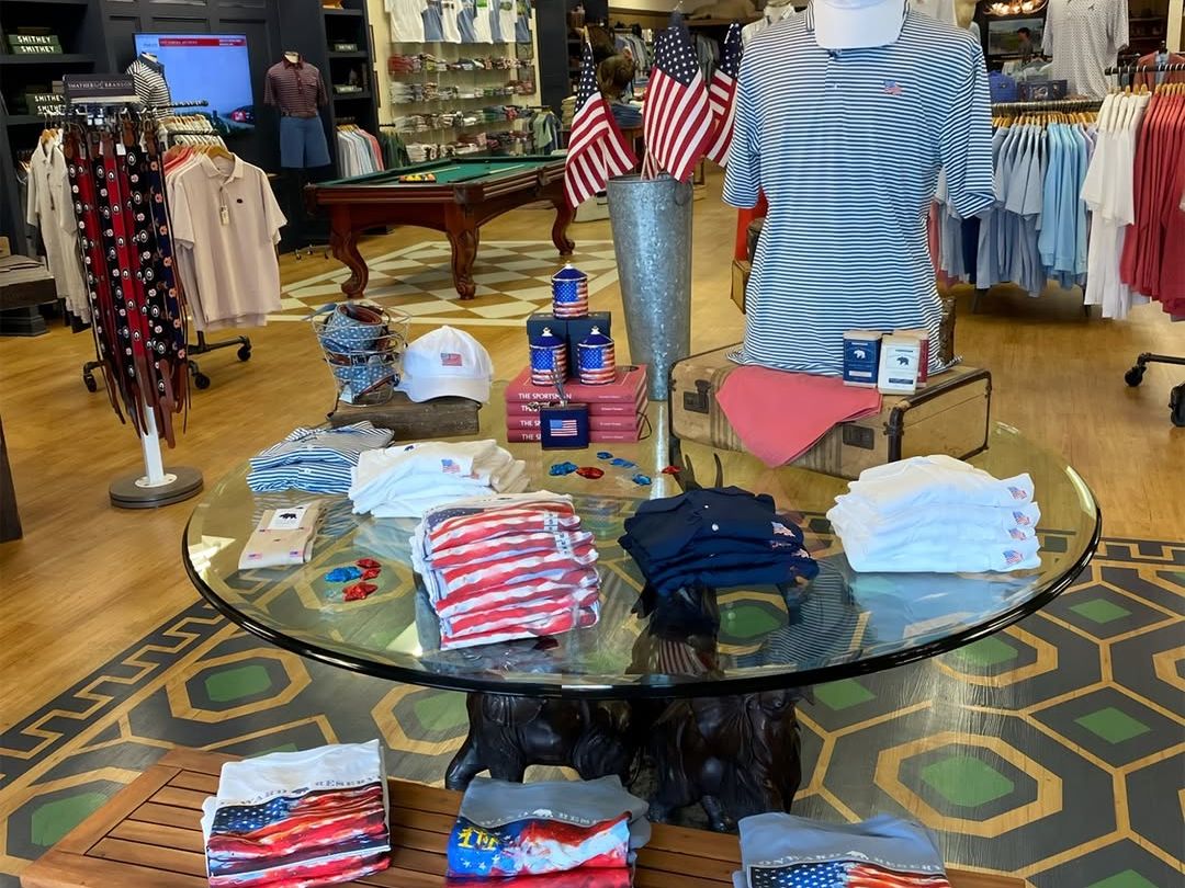 The interior of Onward Reserve displays a curated selection of American-themed apparel and accessories arranged on a round glass-top table with a carved bear base, surrounded by folded flag-print shirts, hats, and koozies. Polo shirts and belts line the walls and racks in the background, with a pool table and mounted deer head adding to the classic Southern sporting aesthetic.