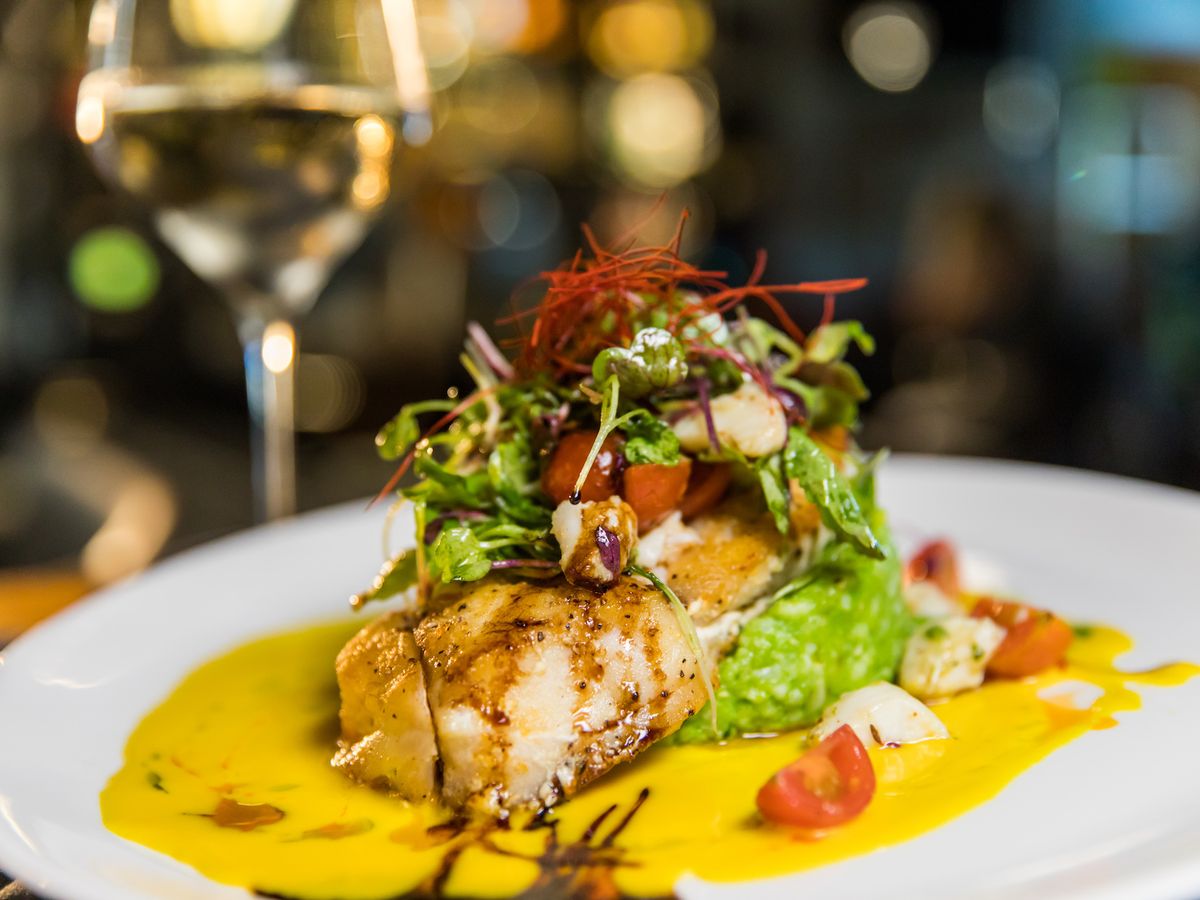 A gourmet dish featuring a perfectly seared fillet of fish on a bed of vibrant green purée, topped with a fresh salad of greens, tomatoes, and nuts. The plate is elegantly finished with a rich yellow sauce and balsamic drizzle, complemented by a glass of white wine in the softly lit background