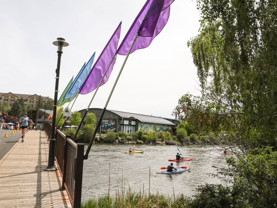 Kayakers paddling along the Deschutes River under colorful flags, with runners crossing a riverside bridge in Bend’s vibrant Old Mill District.