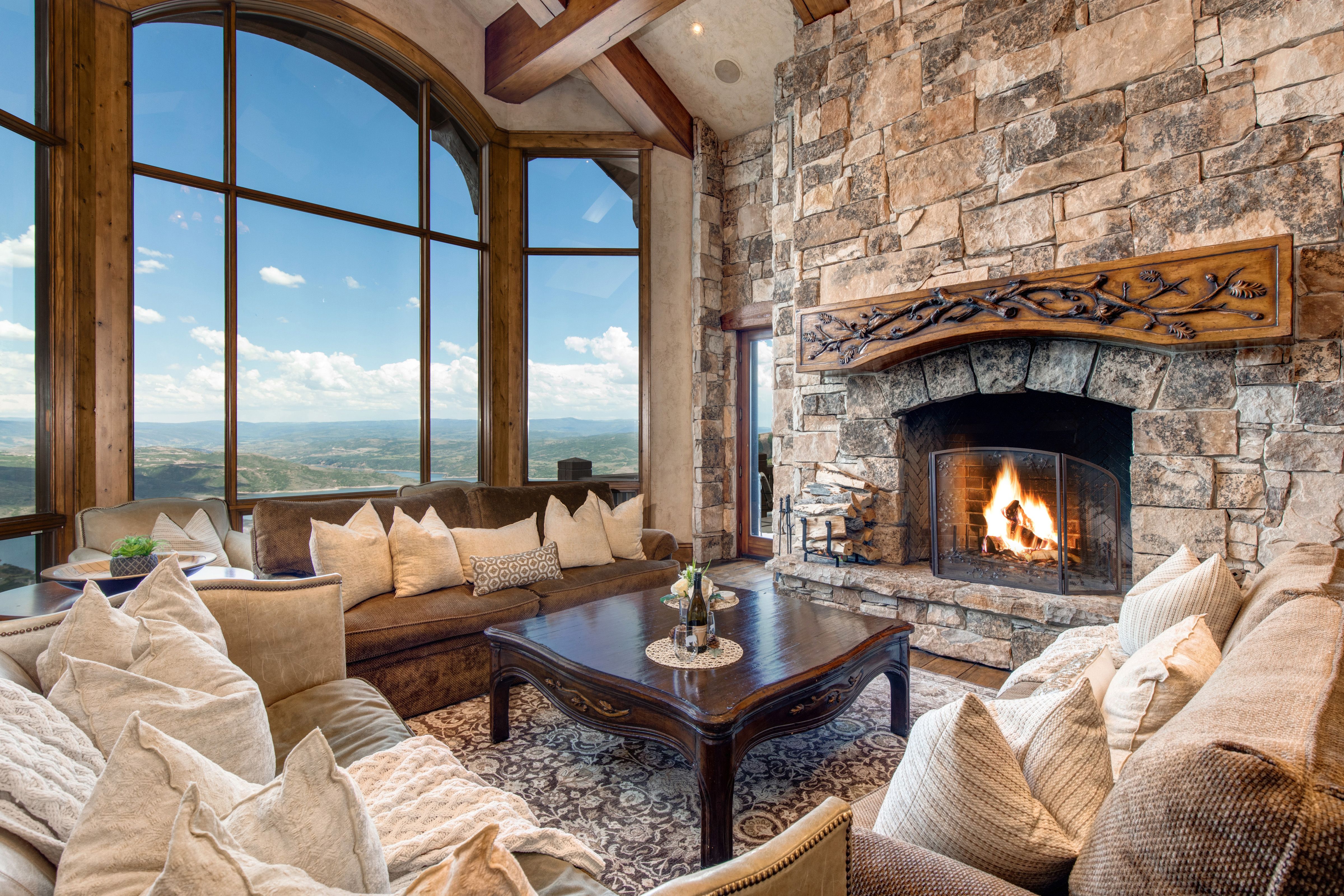A luxurious mountain lodge living room with a grand stone fireplace, plush sofas with neutral-toned pillows, a dark wood coffee table, and floor-to-ceiling arched windows offering sweeping views of the valley and sky.