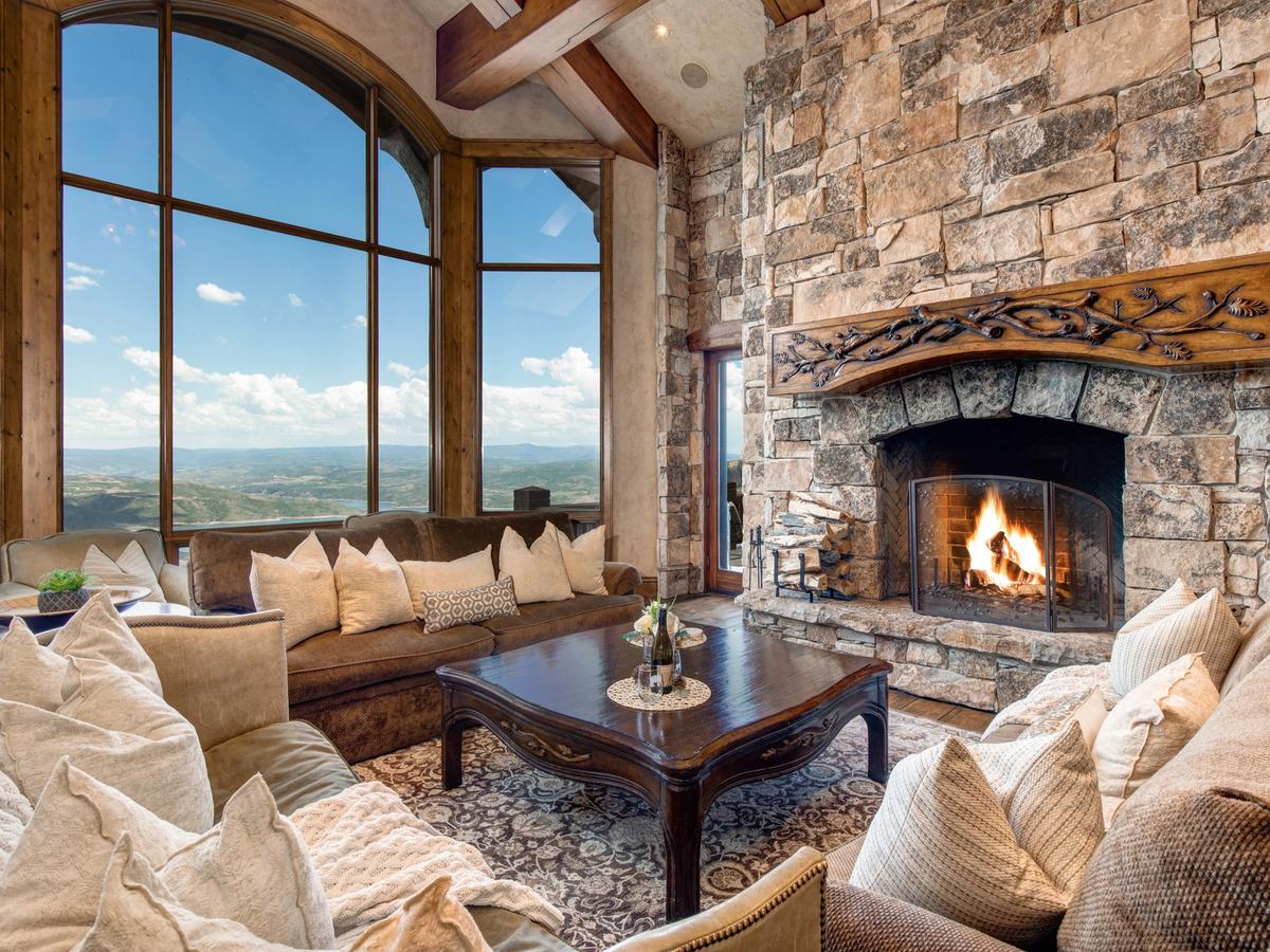A luxurious mountain lodge living room with a grand stone fireplace, plush sofas with neutral-toned pillows, a dark wood coffee table, and floor-to-ceiling arched windows offering sweeping views of the valley and sky.