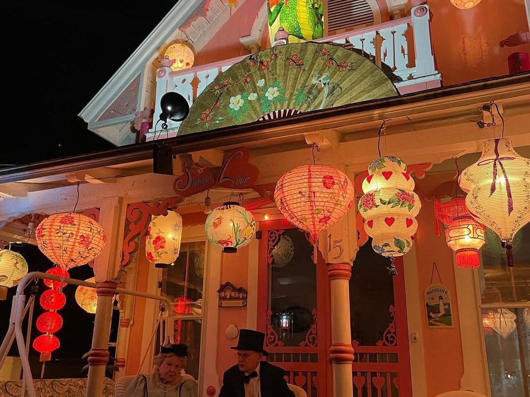 A pastel cottage in Oak Bluffs glows under a canopy of colorful paper lanterns during Grand Illumination Night. Dressed in period clothing, two people sit on the porch, capturing the nostalgic charm and magic of this beloved Martha’s Vineyard tradition.