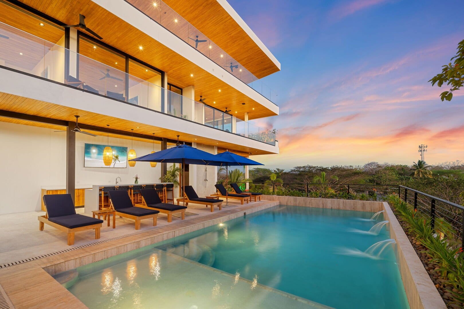 Modern luxury home with a multi-level balcony overlooking an infinity pool, lounge chairs, and umbrellas set against a colorful sunset sky.