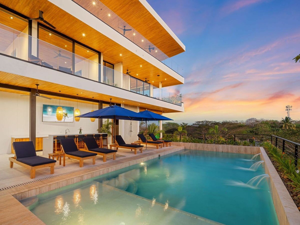 Modern luxury home with a multi-level balcony overlooking an infinity pool, lounge chairs, and umbrellas set against a colorful sunset sky.