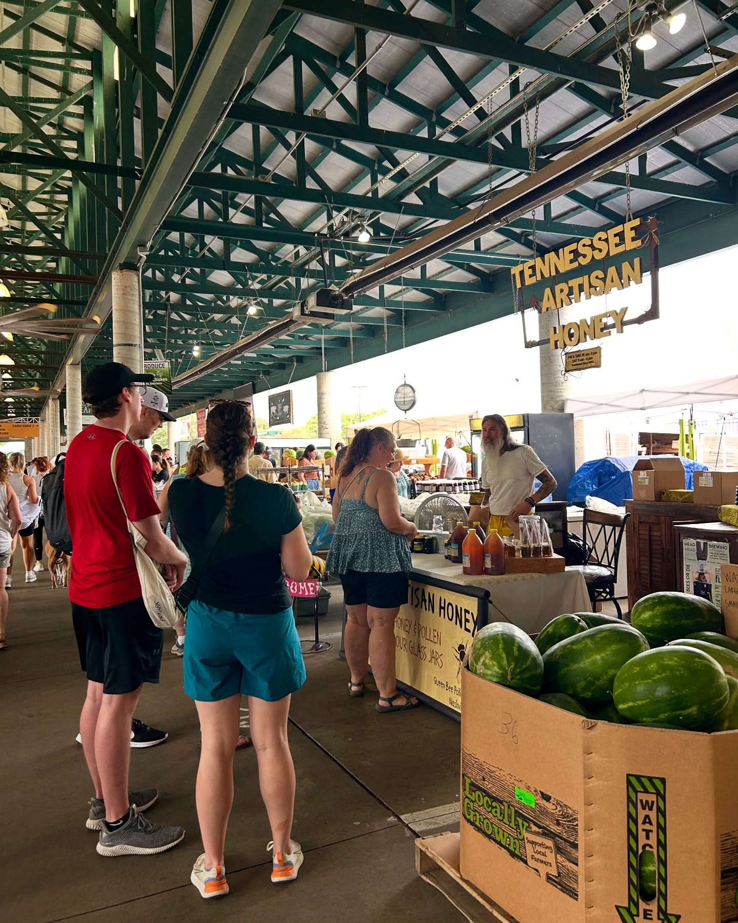 Shoppers explore the Turnip Green Festival inside the Nashville Farmers’ Market, stopping at a booth selling Tennessee artisan honey. Fresh watermelons fill a large box nearby, adding bright color to the busy market scene. The whole moment feels warm and local, with people chatting, tasting, and enjoying the fresh produce.