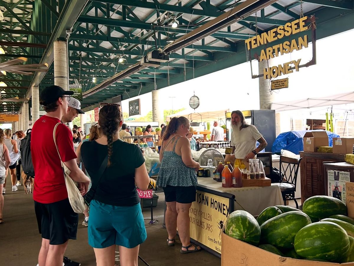 Shoppers explore the Turnip Green Festival inside the Nashville Farmers’ Market, stopping at a booth selling Tennessee artisan honey. Fresh watermelons fill a large box nearby, adding bright color to the busy market scene. The whole moment feels warm and local, with people chatting, tasting, and enjoying the fresh produce.