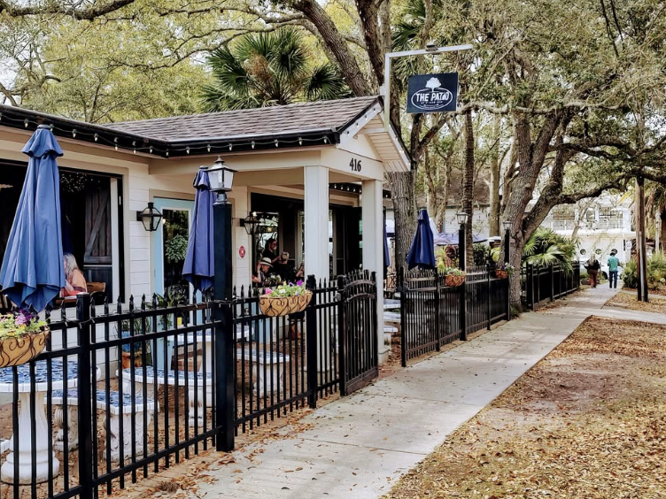 Charming outdoor café, "The Patio," featuring a white cottage-style building with a black fence, blue umbrellas, and stone patio tables. Surrounded by lush trees and a sidewalk, the inviting space offers shaded outdoor seating with hanging flower planters