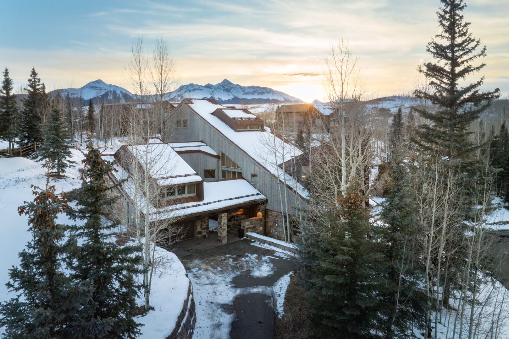 Modern mountain home with snow-covered roof, surrounded by tall trees and a backdrop of snowy peaks at sunset.