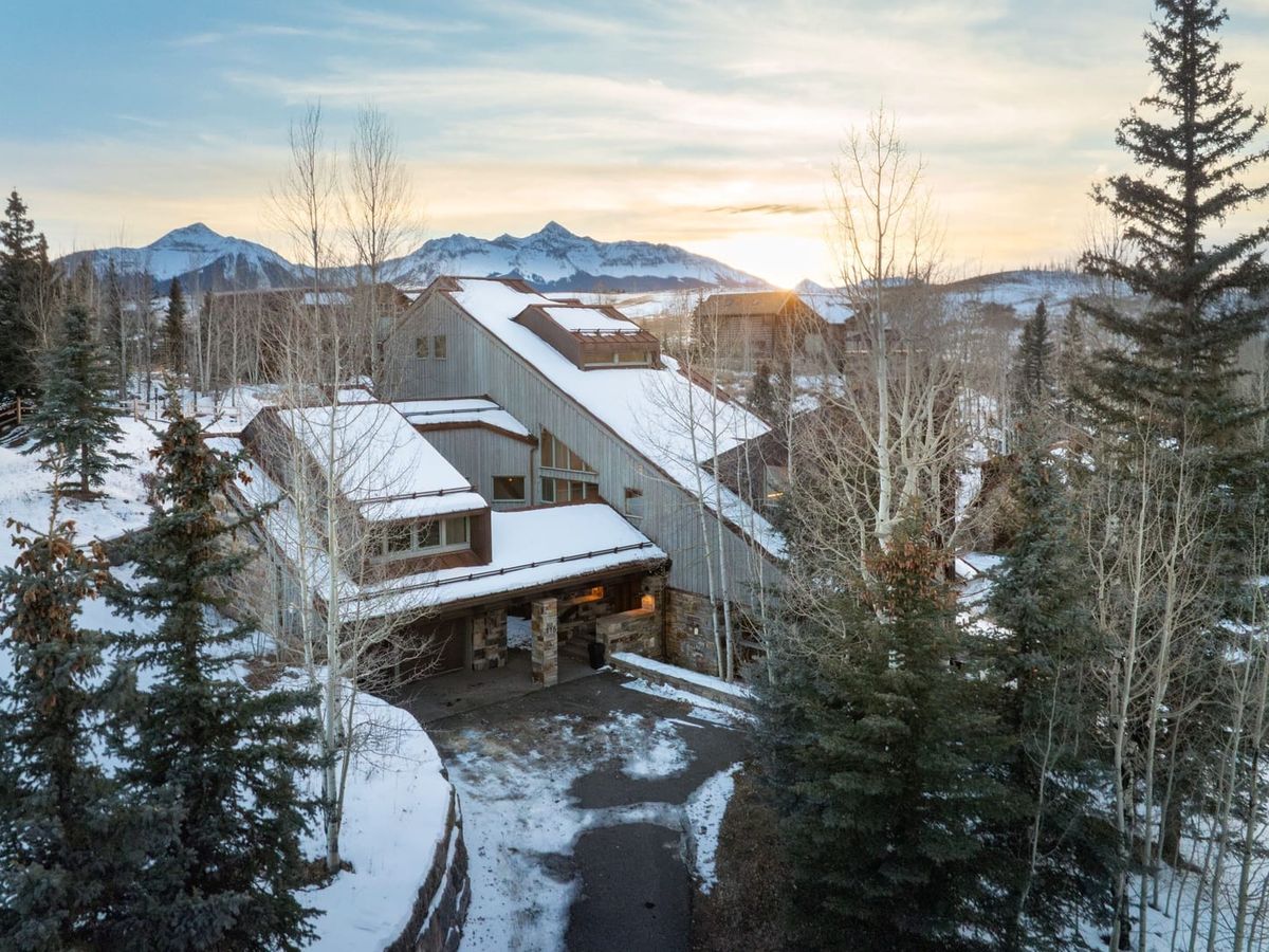 Modern mountain home with snow-covered roof, surrounded by tall trees and a backdrop of snowy peaks at sunset.