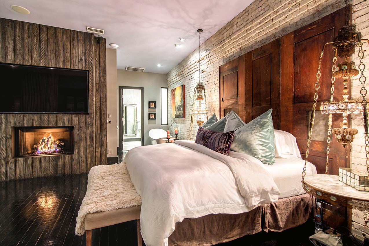 A luxurious, moody bedroom with exposed brick walls, rich wood accents, a plush king bed, and a modern fireplace, blending historic charm with upscale Nashville style.