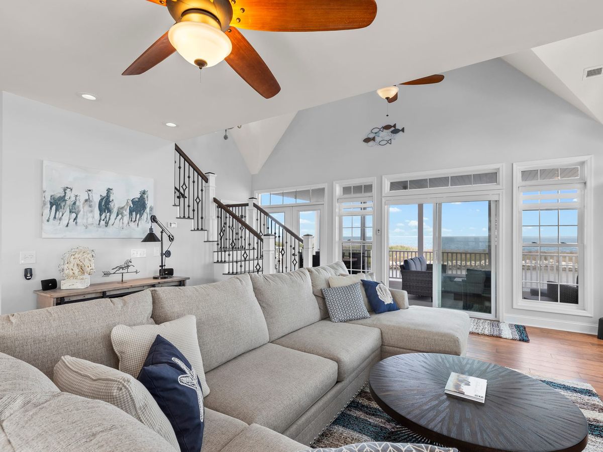 A bright coastal living room with soft gray sofas and big windows looking out over the water. Natural light fills the room, giving it a calm and airy feel. It’s a cozy spot to relax after spending time outside.