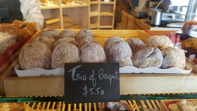 A wooden bakery case displays rows of powdered-sugar-dusted beignets behind glass, with a chalkboard sign reading "Trio of Beignet $5.50." Additional pastries and the warm bakery interior are visible in the background. 