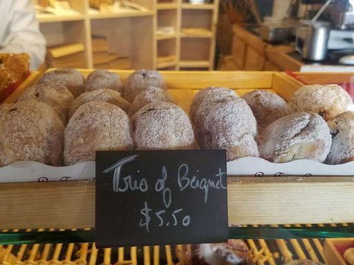 A wooden bakery case displays rows of powdered-sugar-dusted beignets behind glass, with a chalkboard sign reading "Trio of Beignet $5.50." Additional pastries and the warm bakery interior are visible in the background.