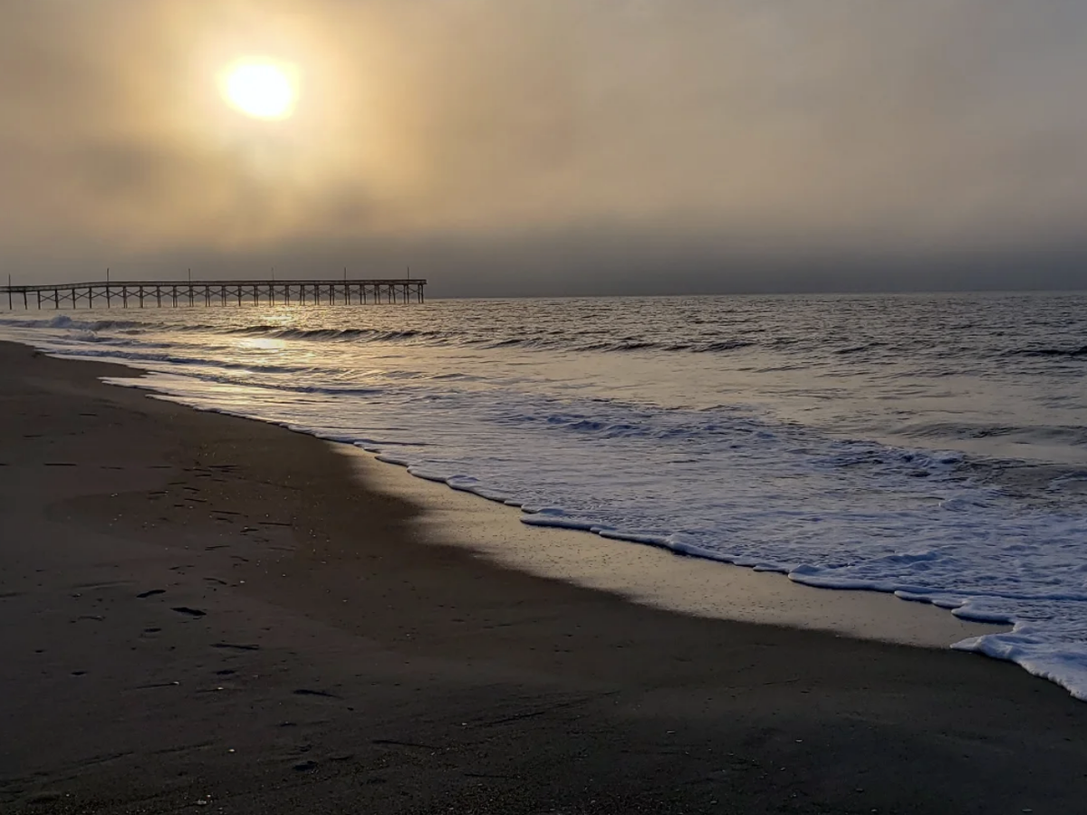 A hazy sunrise casts a golden glow over the ocean as gentle waves wash onto an empty beach with a wooden pier in the distance.