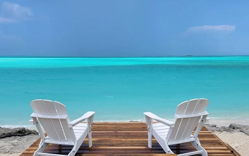 Two white Adirondack chairs sit on a wooden deck facing the calm turquoise ocean, offering a peaceful spot to relax by the shore.