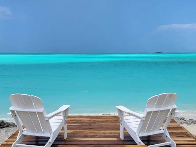 Two white Adirondack chairs sit on a wooden deck facing the calm turquoise ocean, offering a peaceful spot to relax by the shore.