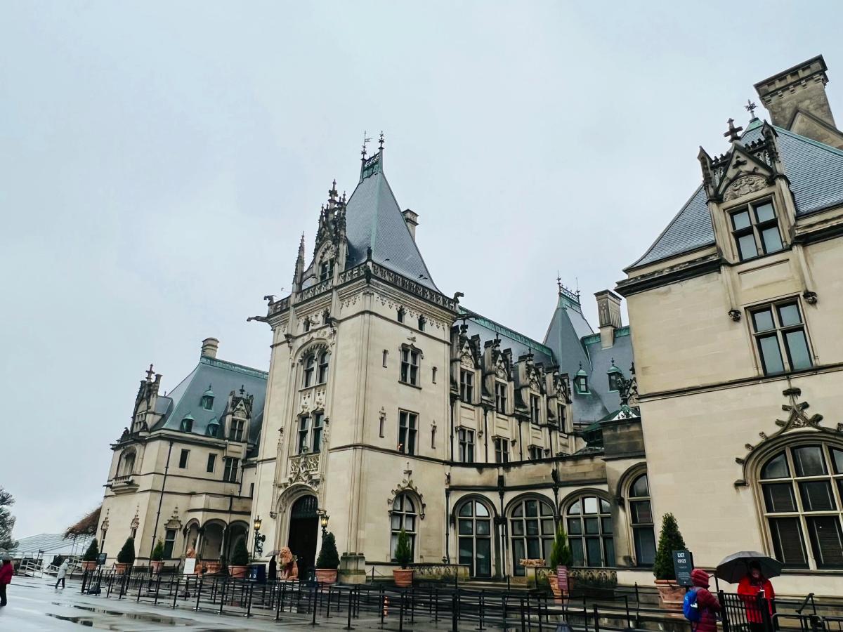 Grand historic mansion with gothic architecture and slate rooftops on a rainy day, with visitors holding umbrellas near the entrance.