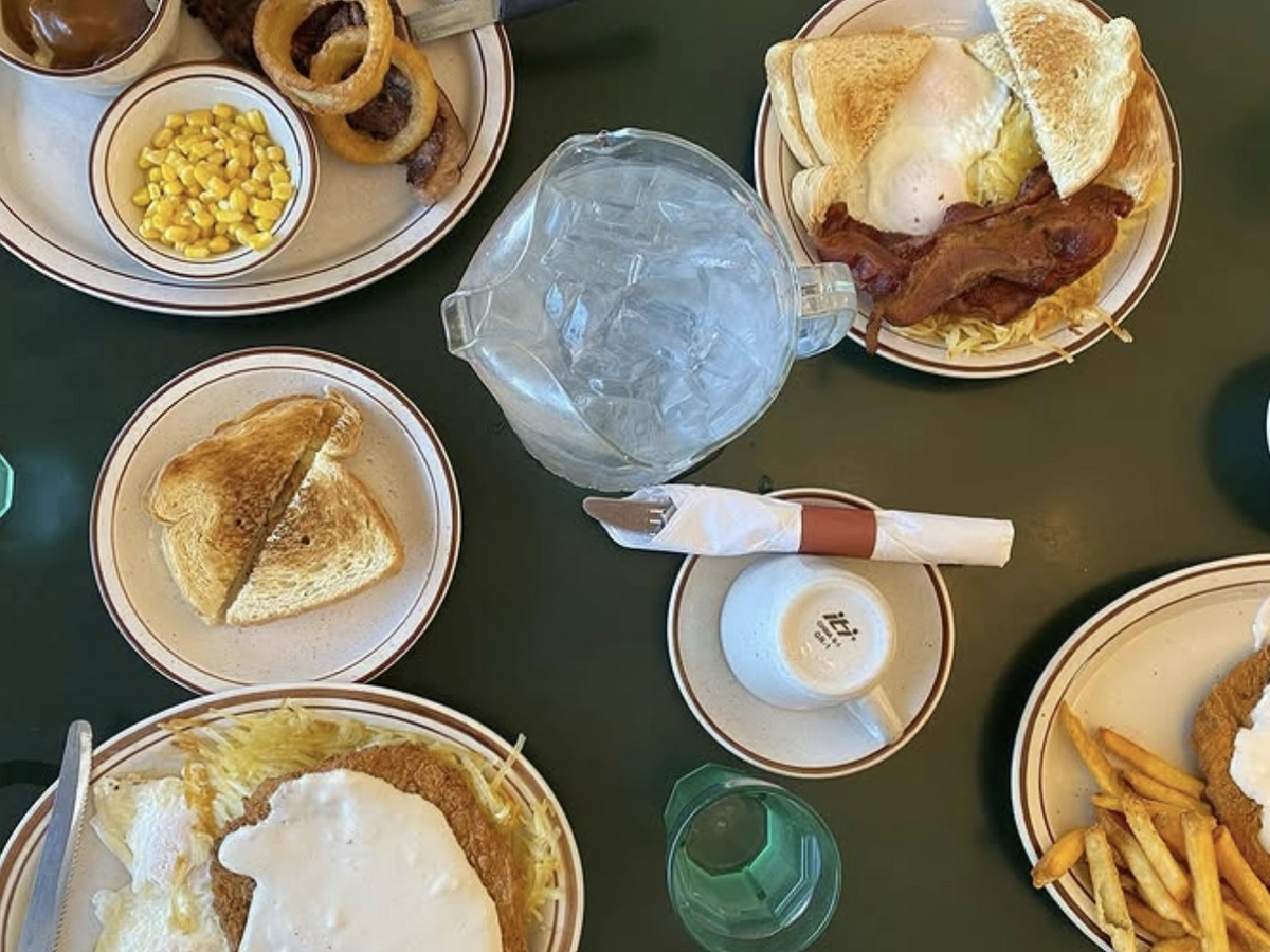Top-down view of a diner-style table set with plates of breakfast and lunch foods including eggs, bacon, toast, hash browns, country fried steak with gravy, fries, corn, onion rings, and a pitcher of ice water.