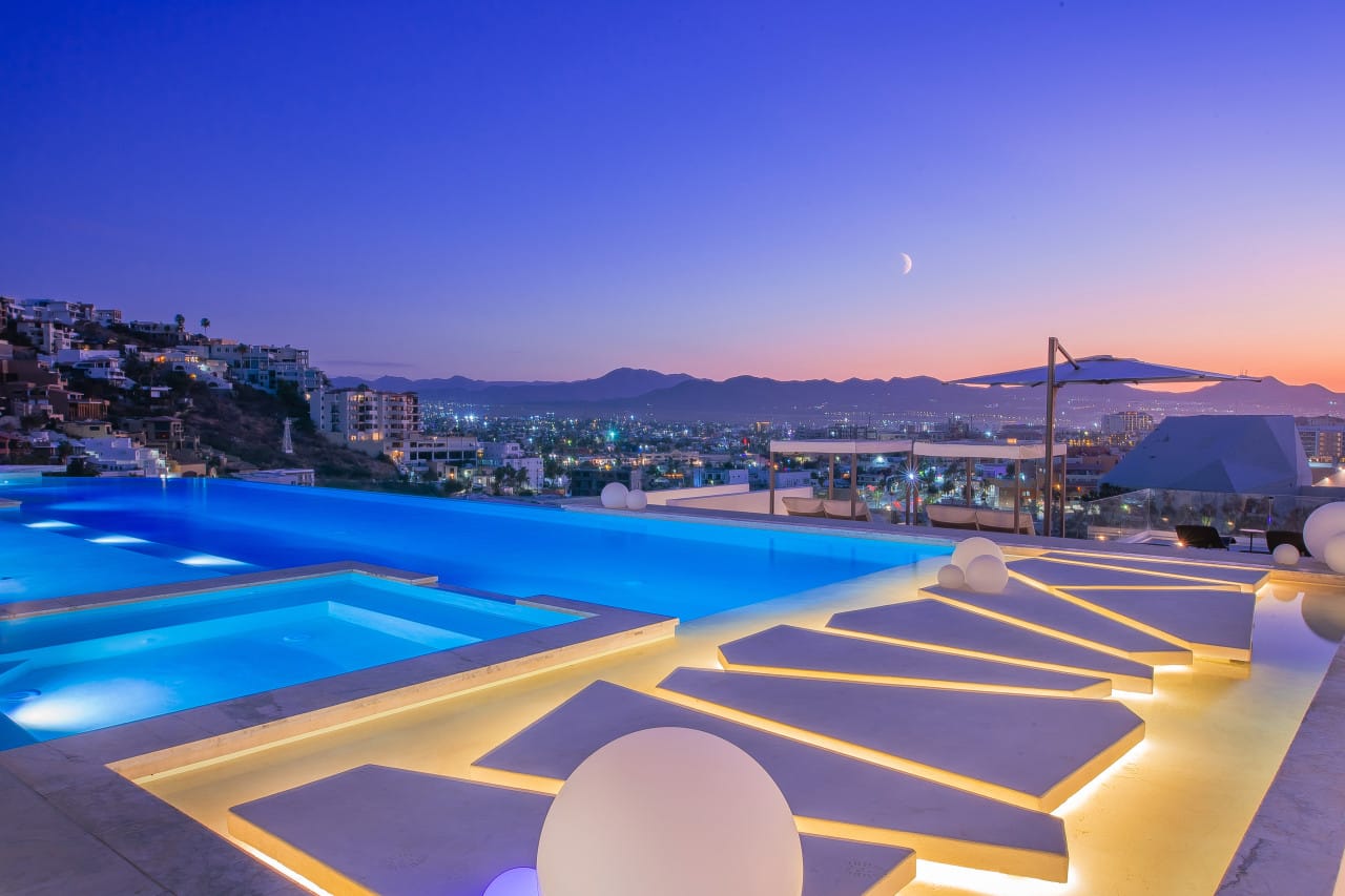Infinity-edge pool and hot tub glowing at dusk with sculptural walkways, modern lounge beds, and panoramic views of a city skyline and surrounding mountains.