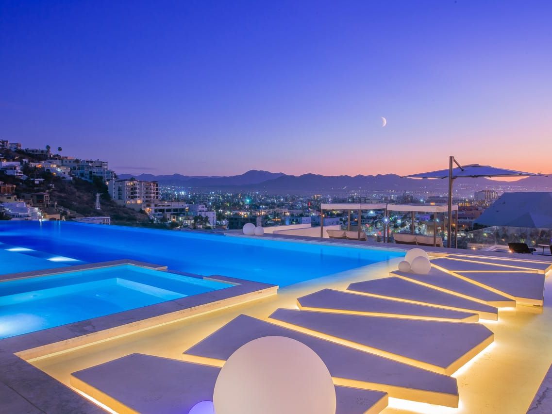 Infinity-edge pool and hot tub glowing at dusk with sculptural walkways, modern lounge beds, and panoramic views of a city skyline and surrounding mountains.
