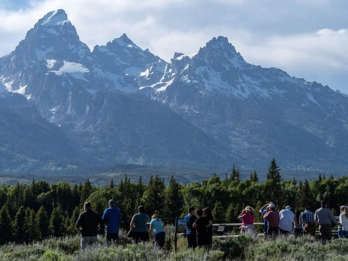 Group of people gathered at a scenic overlook, admiring the snow-capped peaks of a dramatic mountain range under a partly cloudy sky.