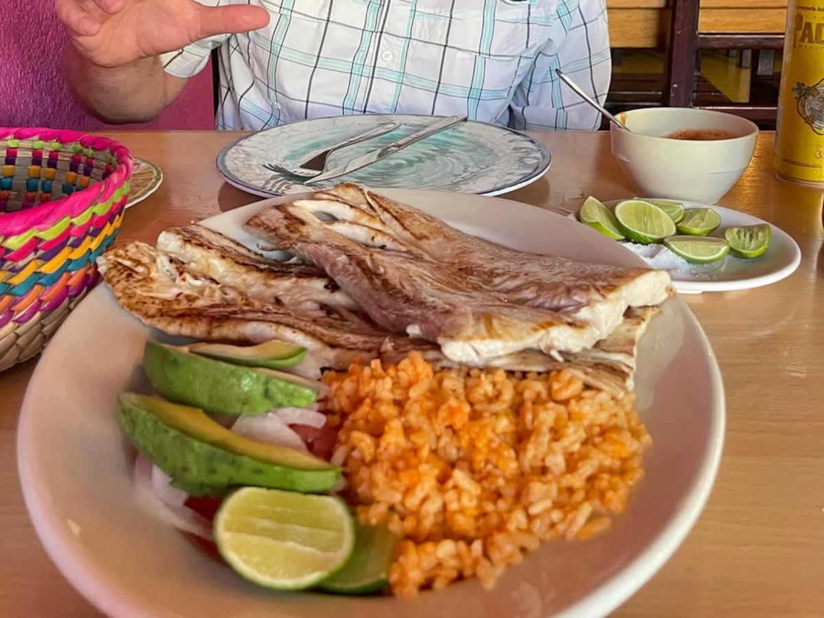 A plate of grilled fish with avocado, lime wedges, and a side of seasoned rice, accompanied by a basket, a small bowl of sauce, and a beverage in the background.