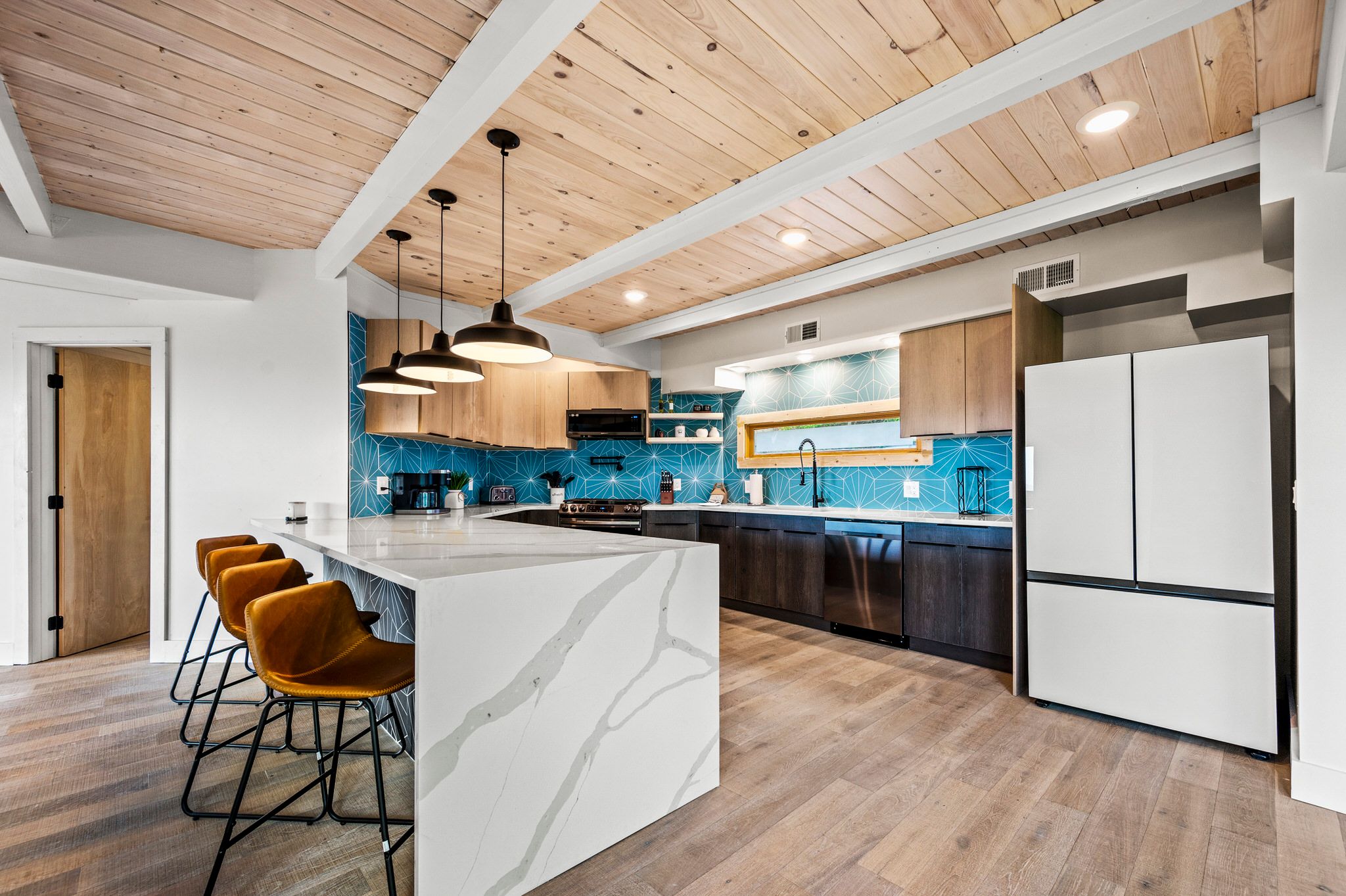 A stylish modern kitchen with a white marble waterfall island, mustard leather barstools, wood-paneled ceiling, and striking blue geometric tile backsplash paired with light and dark wood cabinetry.