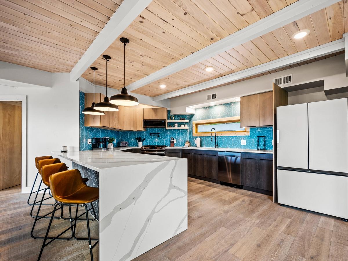 A stylish modern kitchen with a white marble waterfall island, mustard leather barstools, wood-paneled ceiling, and striking blue geometric tile backsplash paired with light and dark wood cabinetry.