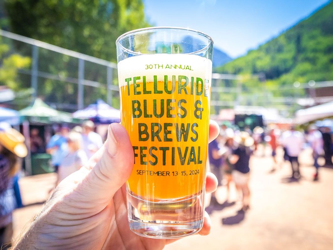 A cold craft beer held up at the Telluride Blues & Brews Festival, with a crowd and tents blurred in the background. This lively fall festival mixes great beer, mountain air, and live music.