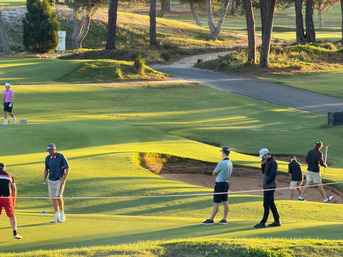 A group of golfers spread out across a well-bunkered fairway at golden hour, studying their shots near a sand trap with tall pine trees and winding cart paths visible in the background.