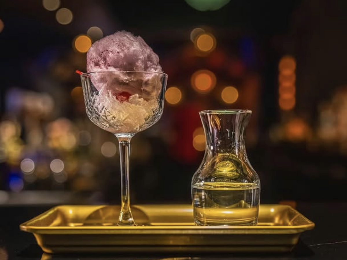 A craft cocktail served in a crystal coupe glass with cotton candy and ice, paired with a small carafe of liquid on a golden tray against a blurred bar backdrop.