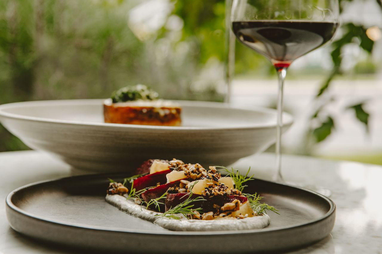 A beautifully styled plate features roasted vegetables, herbs, nuts, and a smooth sauce, paired with a glass of red wine. The soft, natural light gives the meal a calm and elegant feel. It highlights the seasonal, upscale style of Ariana Restaurant