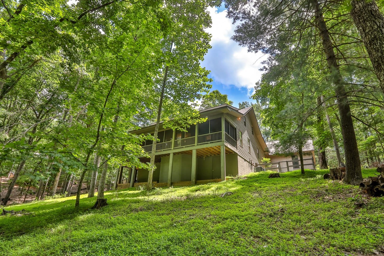 Cozy mountain cabin exterior set on a green hillside surrounded by tall trees and forest views. The peaceful wooded setting makes it a great choice for guests looking for privacy and a relaxing mountain getaway.