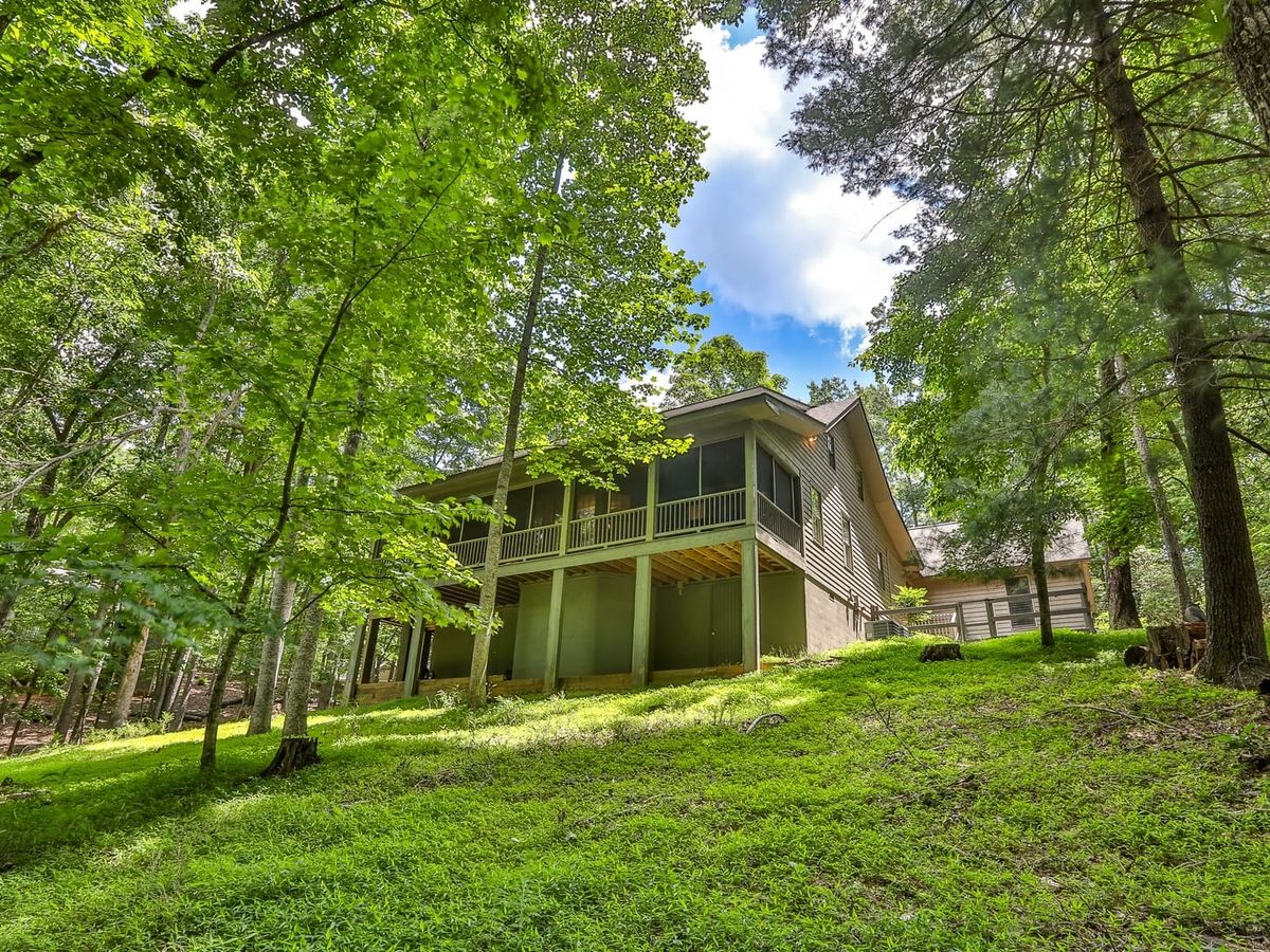 Cozy mountain cabin exterior set on a green hillside surrounded by tall trees and forest views. The peaceful wooded setting makes it a great choice for guests looking for privacy and a relaxing mountain getaway.