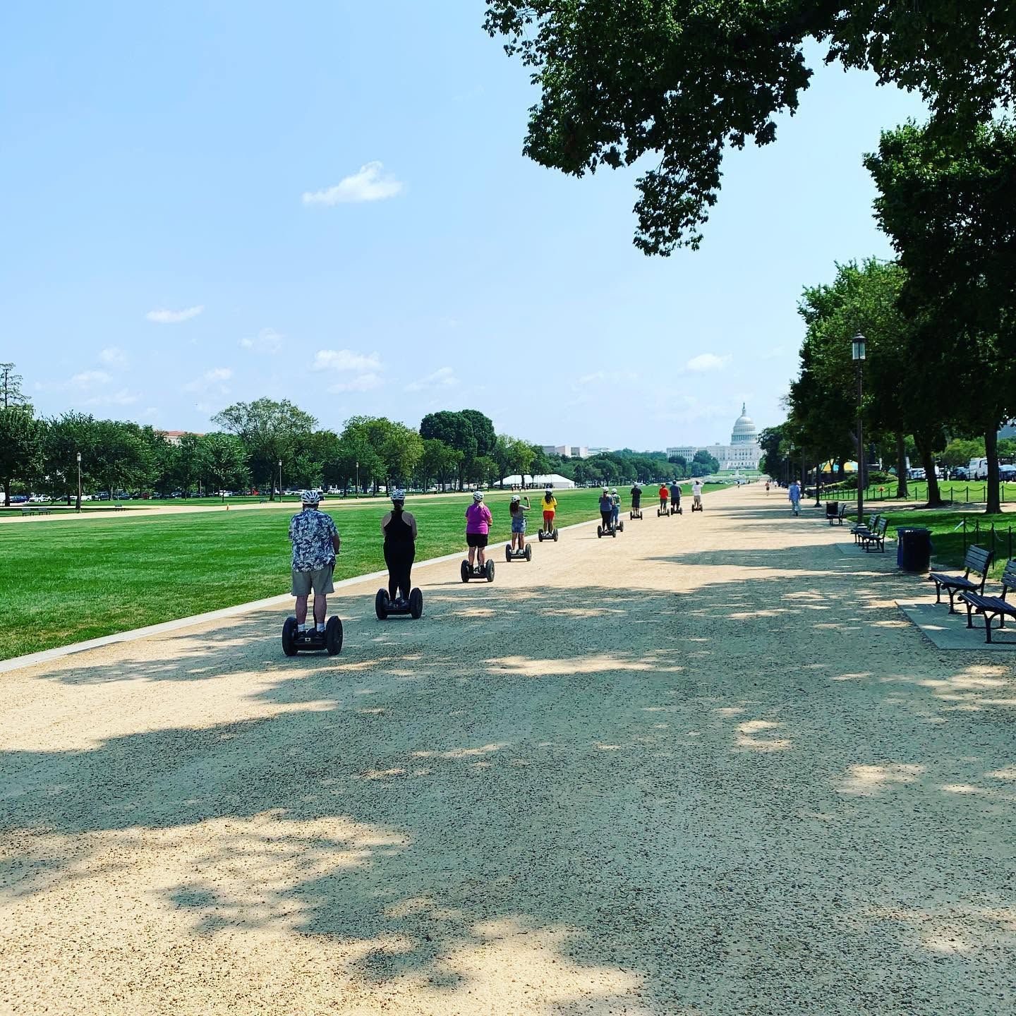 A group of tourists glide along the gravel path of the National Mall on Segways with Capital Segway, heading toward the iconic dome of the US Capitol building under a clear blue summer sky. 